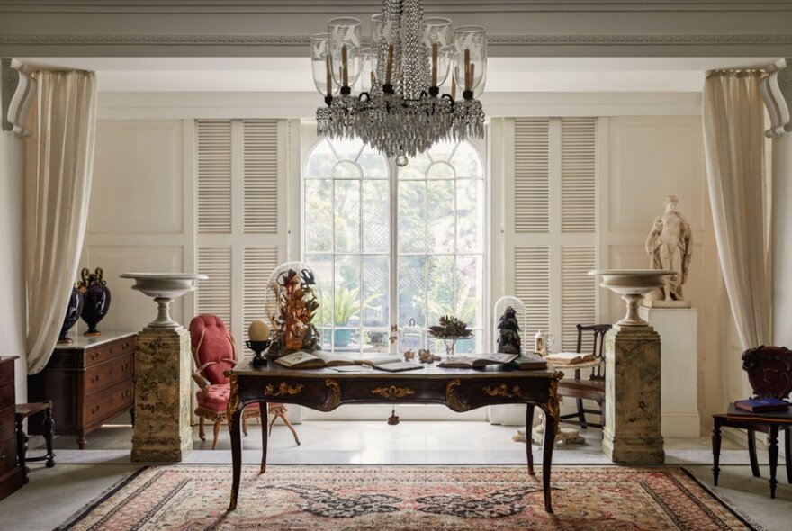 A light-filled Georgian style formal room with a central table, armchairs by the arched window, rugs, statues and other ornate objects and furnishings.