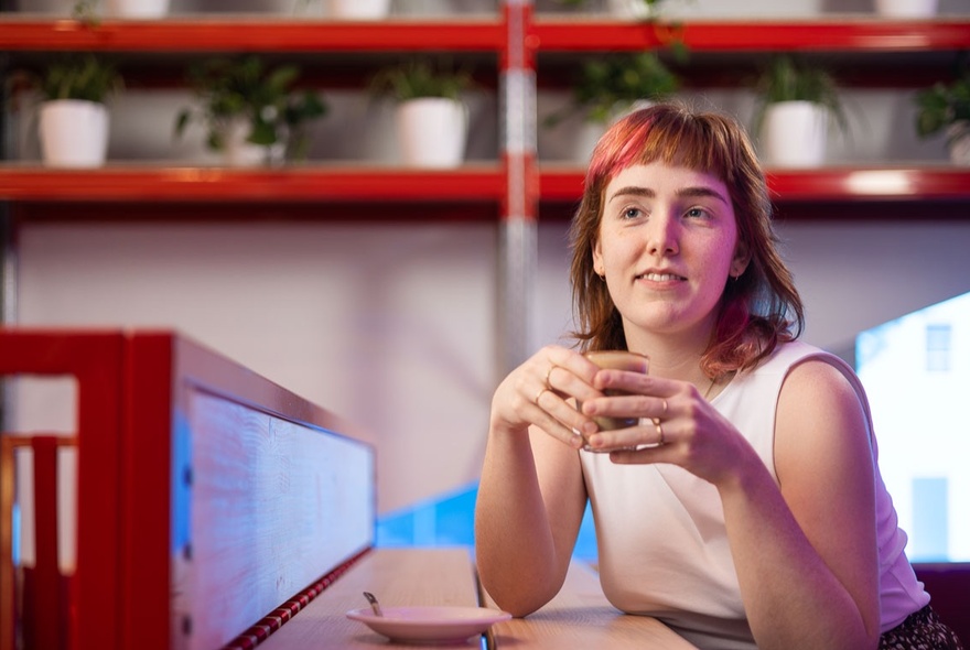 A woman holding a cup of coffee and smiling, seated at a high table, inside the Science Gallery cafe.