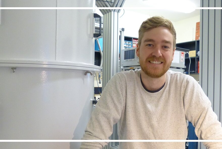 Dr Ben McAllister, a young man with a beard and wearing a white top, smiling while standing next to a large white object.
