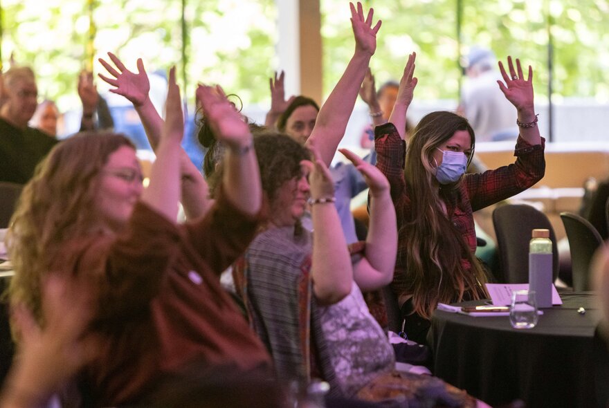 Women in a conference setting, ohlding their arms in the air while seated.