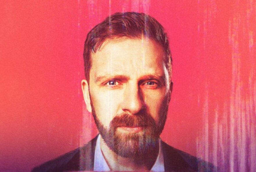 Comedian Pierre Novellie, a man with short red-brown hair and a beard, looking intently at the viewer, posed against a red background