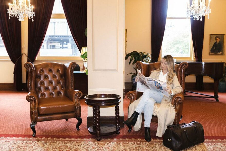 Person reading a broadsheet newspaper in a leather armchair in the lobby of The Windsor, , with chandeliers, heavy velvet drapes and a Persian rug surrounding.