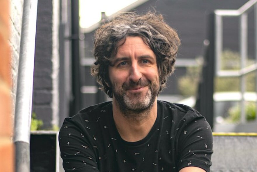 Comedian Mark Watson, a middle-aged man with wavy, slightly messy hair and a beard, smiling slightly and wearing a dark t-shirt; seated outdoors. 