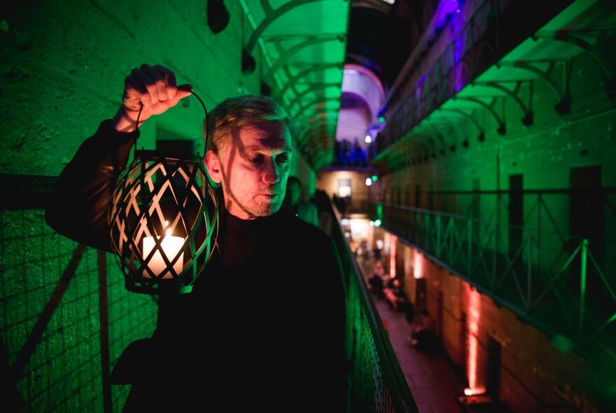 A person holding a lantern near their face inside the Old Melbourne Gaol, dimly lit in purple and green. 