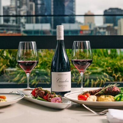 Testaurant table set for dinner with meat platters, wine glasses and wine, with trees and city buildings in the background.
