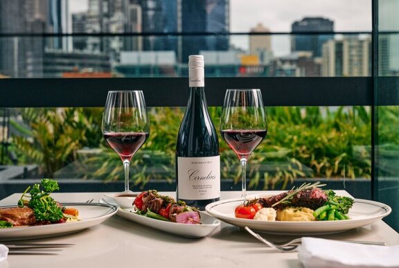 Testaurant table set for dinner with meat platters, wine glasses and wine, with trees and city buildings in the background.