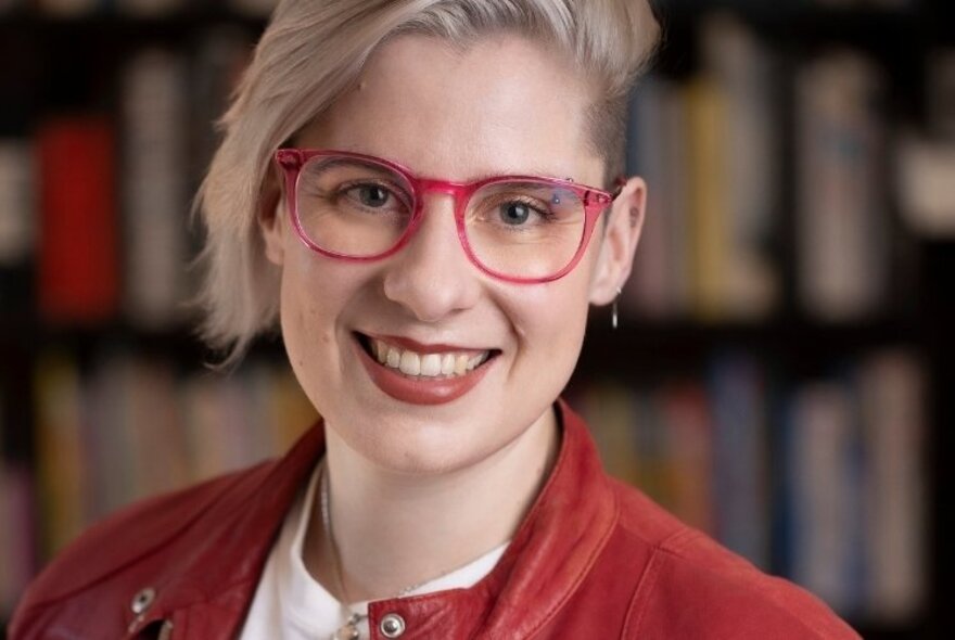 Young person with red framed glasses and short blond hair, wearing a white shirt and red jacket.
