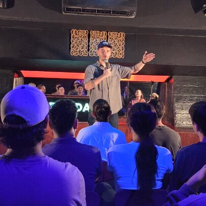 A person on a stage, speaking to a seated live audience at a venue hosting stand-up comedy.