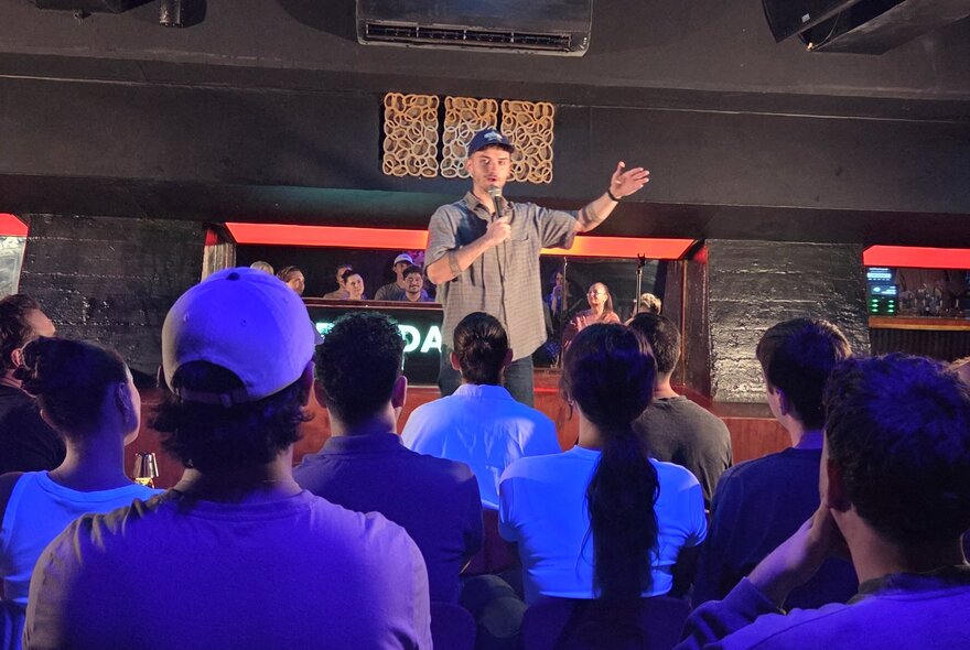 A person on a stage, speaking to a seated live audience at a venue hosting stand-up comedy.