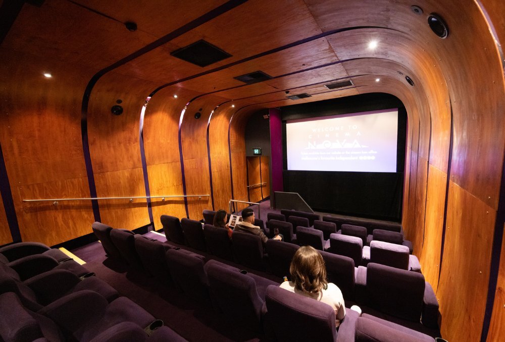 Three people sitting in a movie theatre with purple seats and wooden walls.