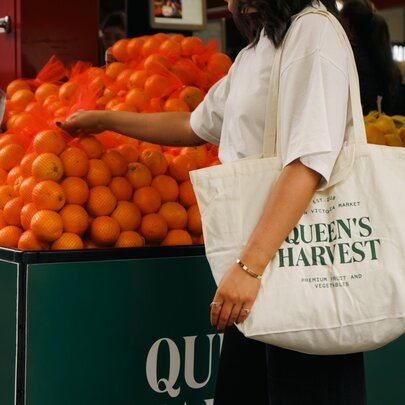 A woman (no head visible) carrying a Queen's Harvest tote bag over her shoulder touching a pyramid of oranges at a market stall. 