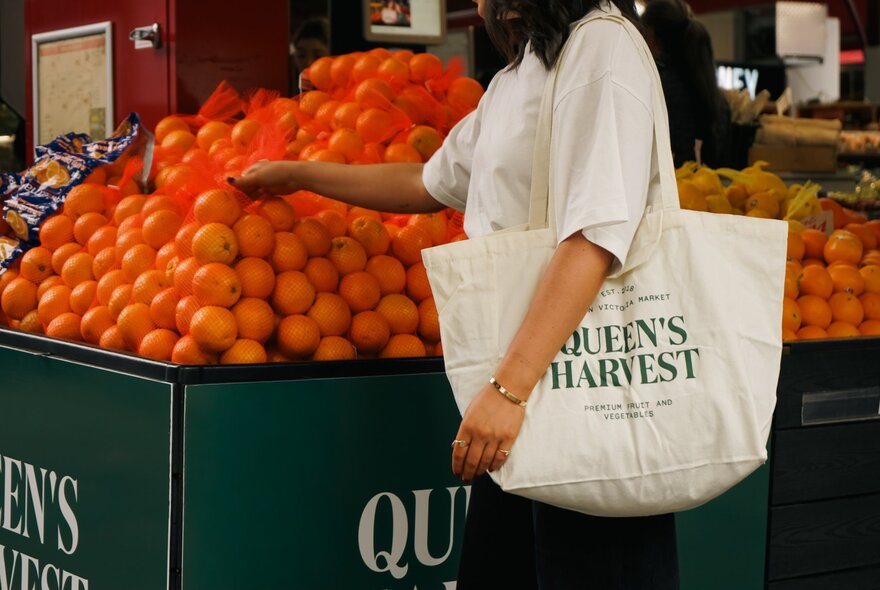 A woman (no head visible) carrying a Queen's Harvest tote bag over her shoulder touching a pyramid of oranges at a market stall. 