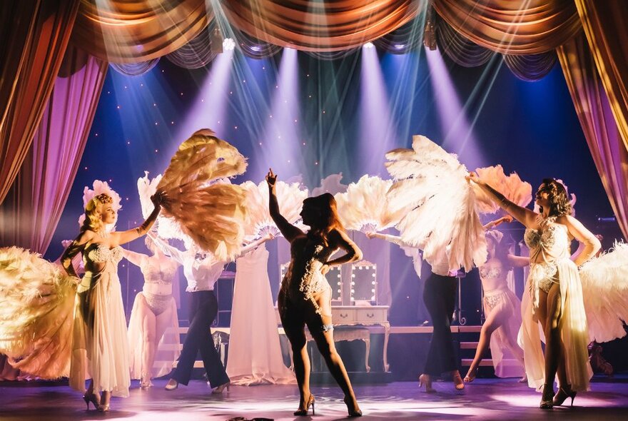 Burlesque performers on a stage in the production of Shimmery Burlesque, wearing costumes and holding oversized feathered fans.