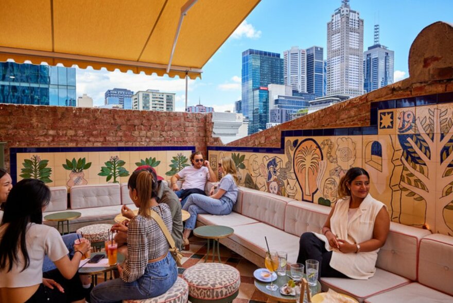 People sitting on couches in a city rooftop bar with tiled wall murals.