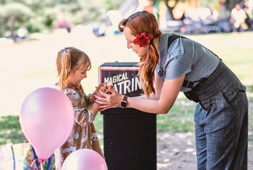Performer Magical Katrina interacting at an outdoor show with a young child from the audience, pink balloons in the foreground.