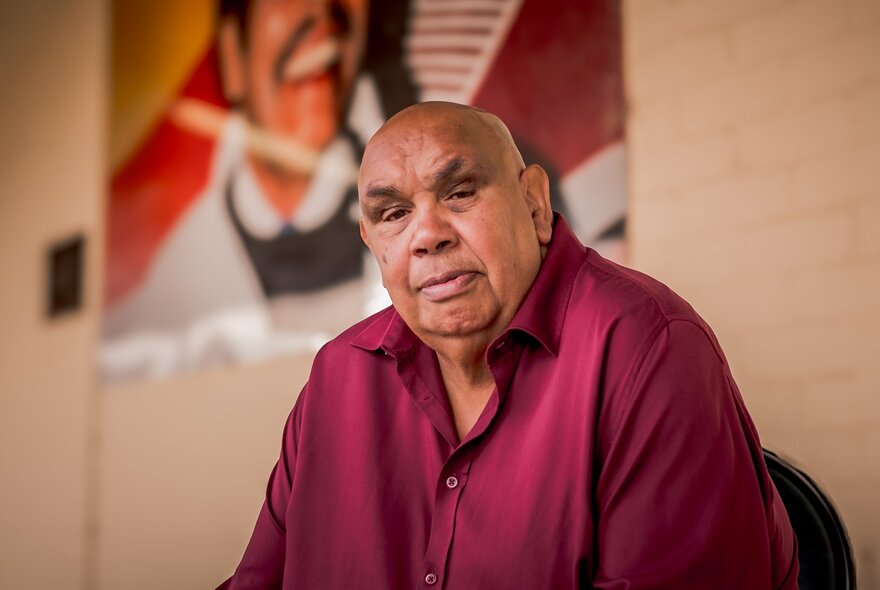 Indigenous musician Kutcha Edwards, seated beneath a poster, wearing a maroon shirt.