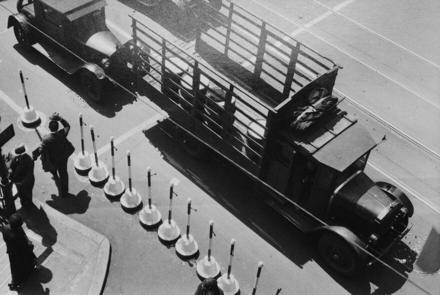 Vintage view looking down onto a parked truck and car next to traffic bollards, with men walking past.