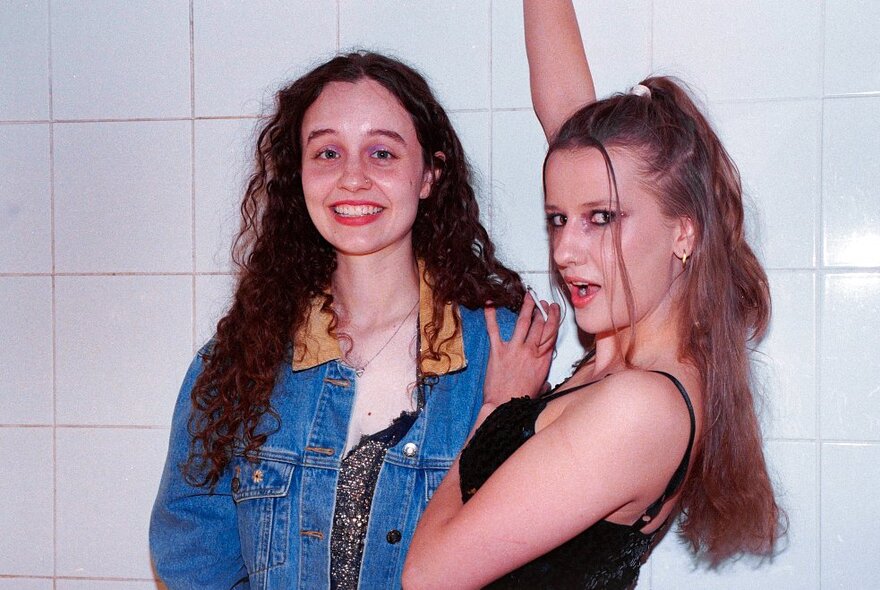 Two girls standing together laughing against a white tiled wall, one with her arm raised above her head.