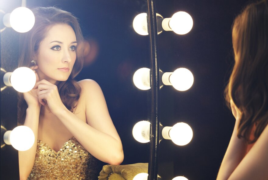 Internationally renowned soprano, Claire Lyon, seen as a reflection in a brightly lit mirror in a theatre dressing room, wearing a gold sequined dress.