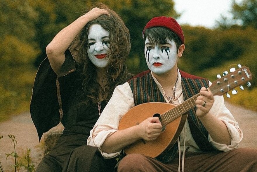 A couple wearing white makeup with black tears, one playing a mandolin, seated outside.