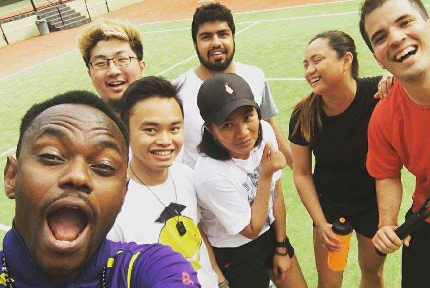A group of smiling people standing on a tennis court, some holding racquets, taking a selfie