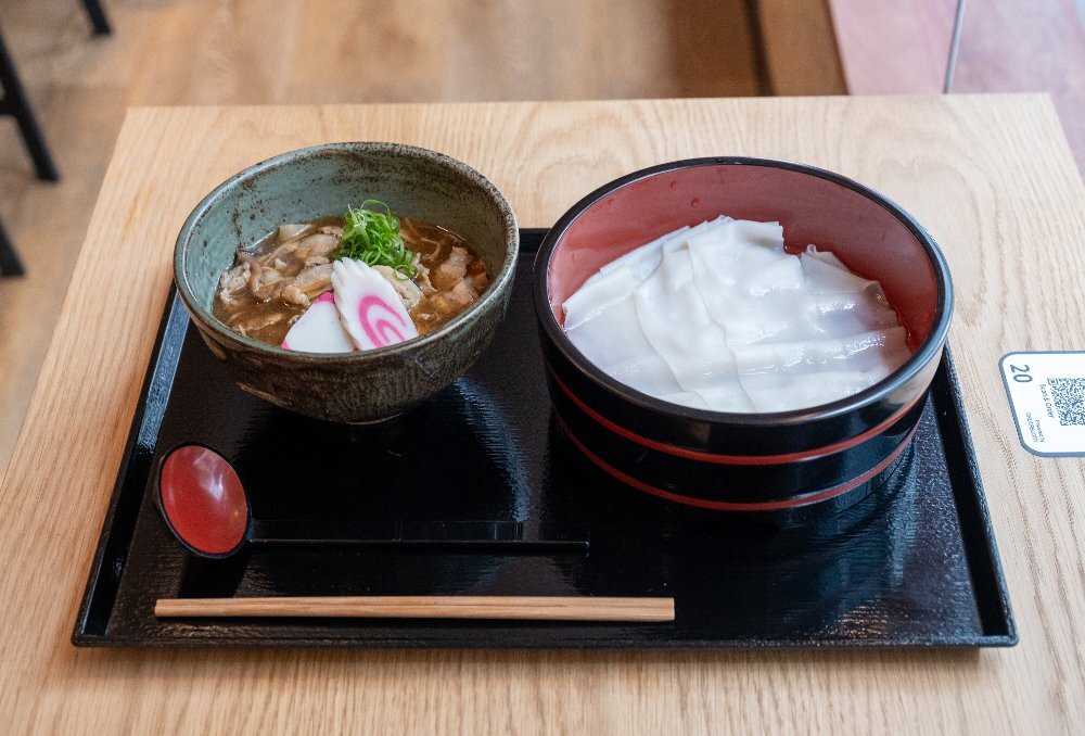 A black serving plate with two bowls: one with a beef broth and the other with layers of wide Himokawa udon noodles.