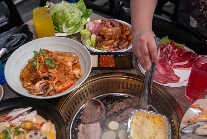 A person is cooking Korean bbq. There are plates of meat, salad and seafood.