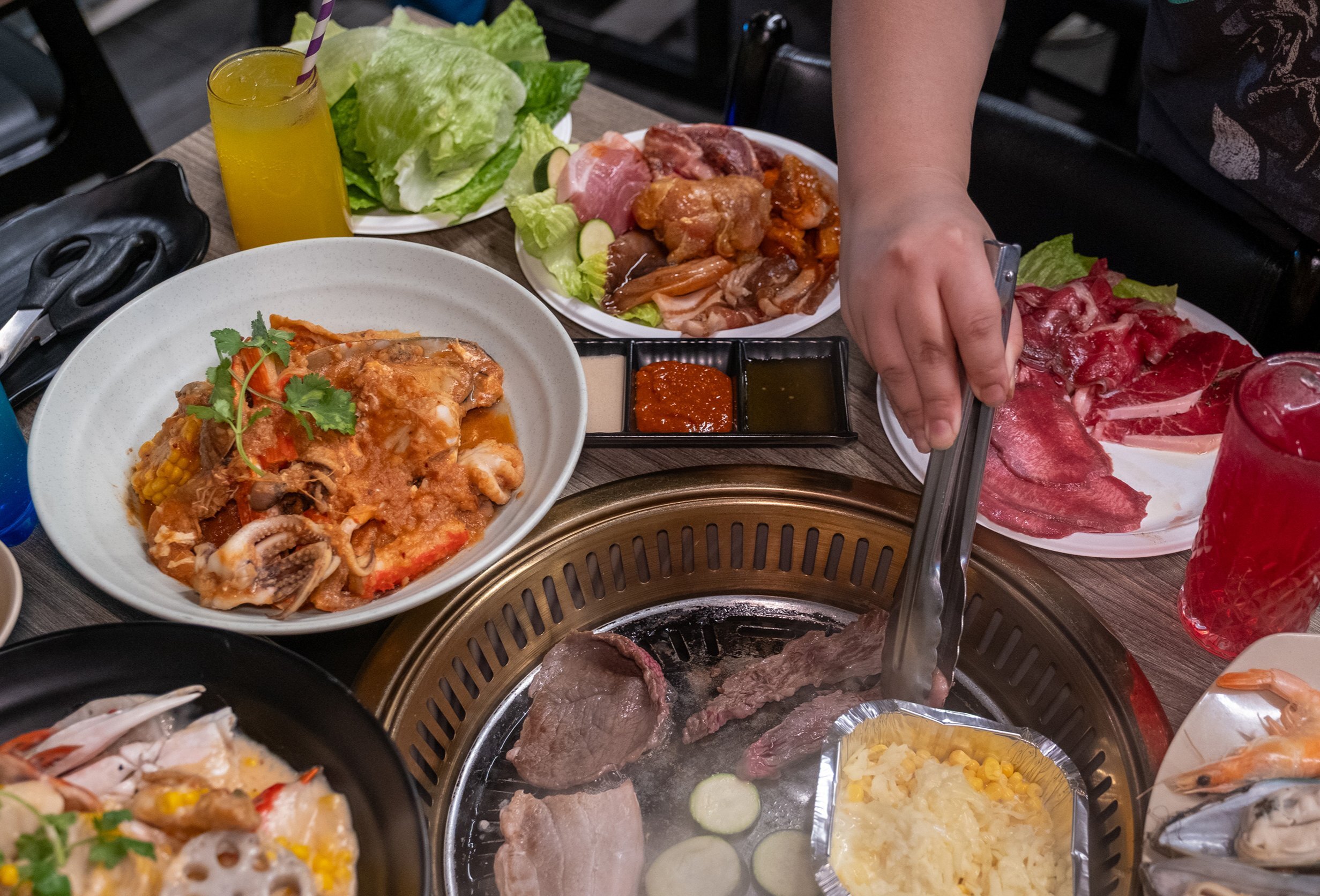 A person is cooking Korean bbq. There are plates of meat, salad and seafood. 