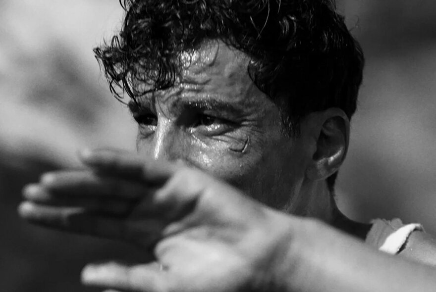 Black and white still from a film showing a man holding his hand up and squinting into the sun.
