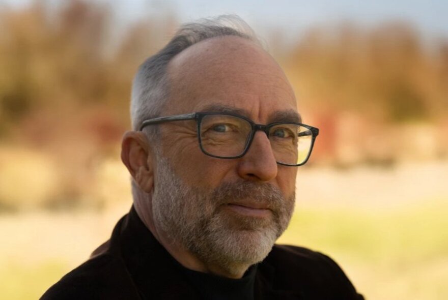 Wikipedia founder, Jimmy Wales, slightly smiling for a portrait with a bright but blurred lanscape in the background.