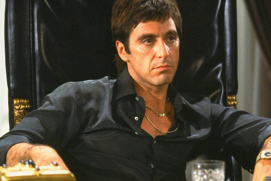 A scene from the film Scarface with the actor Al Pacino as the character Tony Montana, wearing a black shirt he sits in a black leather chair.