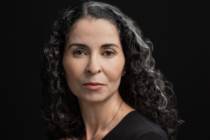 Portrait of Moroccan American writer, Laila Lalami, as she looks seriously at the camera, her curly black and grey hair falling down her back. 