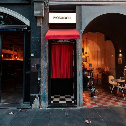 Vintage-style photo booth with red curtains and check floor, next to restaurant interior with similar design and arched windows.