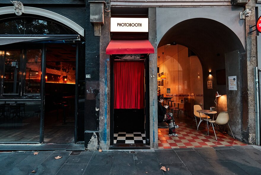 Vintage-style photo booth with red curtains and check floor, next to restaurant interior with similar design and arched windows.