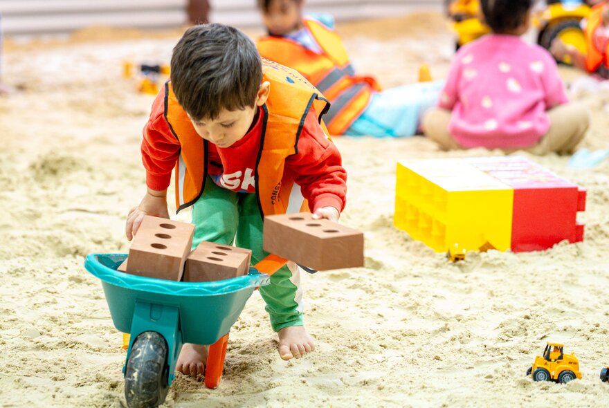 A child playing in a large indoor sandpit using a mini-wheelbaroow filled with foam bricks.