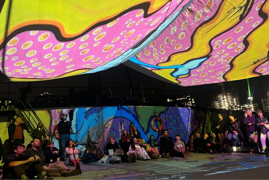 A crowd of people, some seated on the ground, watching a digital projection.