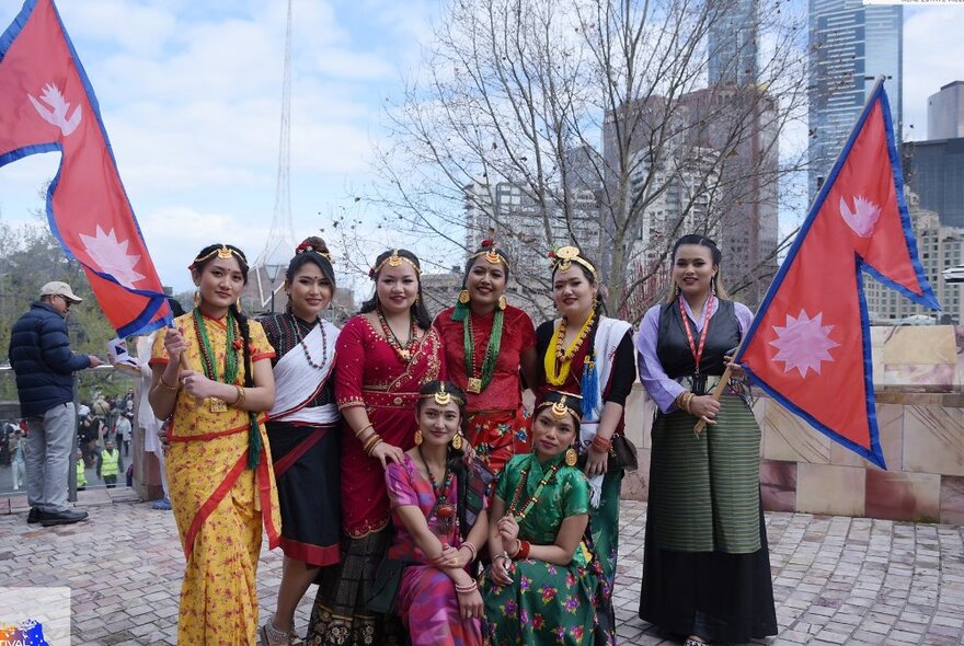 A group of women dressed in traditional and colourful Nepalese costumes, two of them holding flags, in an open square.