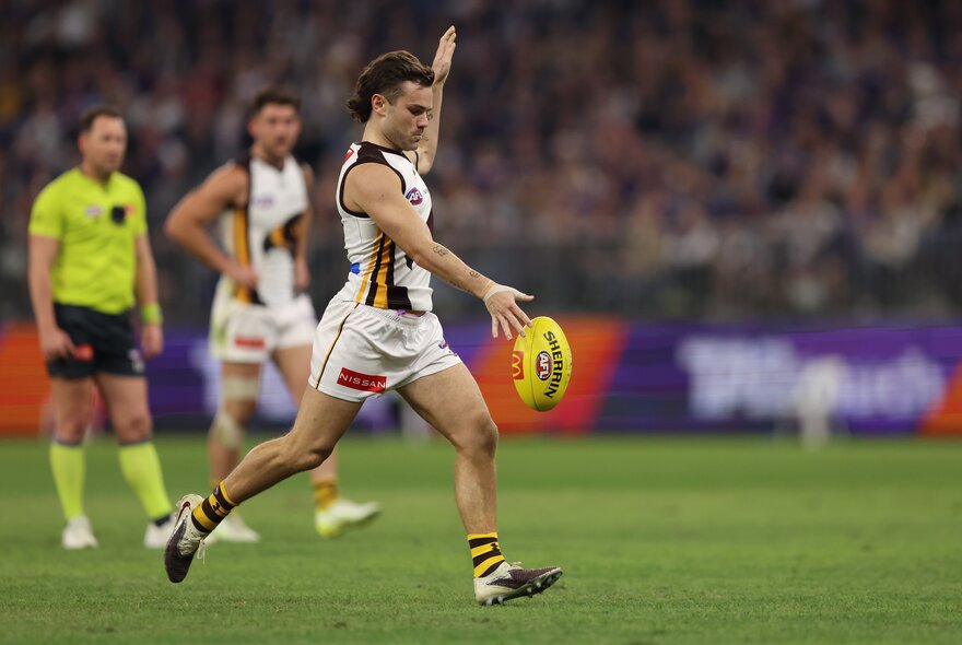 A Hawthorn AFL football player about to kick the football on the field during a match, a blurred crowd of stadium fans in the background, along with an umpire and another player.