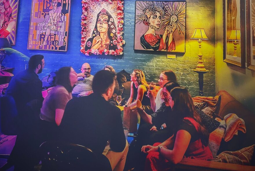 People socialising and drinking in a bar on couches, bright artworks hanging on the blue wall.