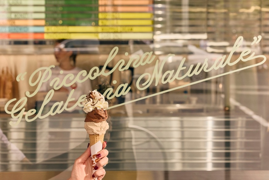A hand holding up an ice cream with two scoops, in front of a shop window that has the words 'Piccolina Gelateria Naturale' written in cursive script on the window.