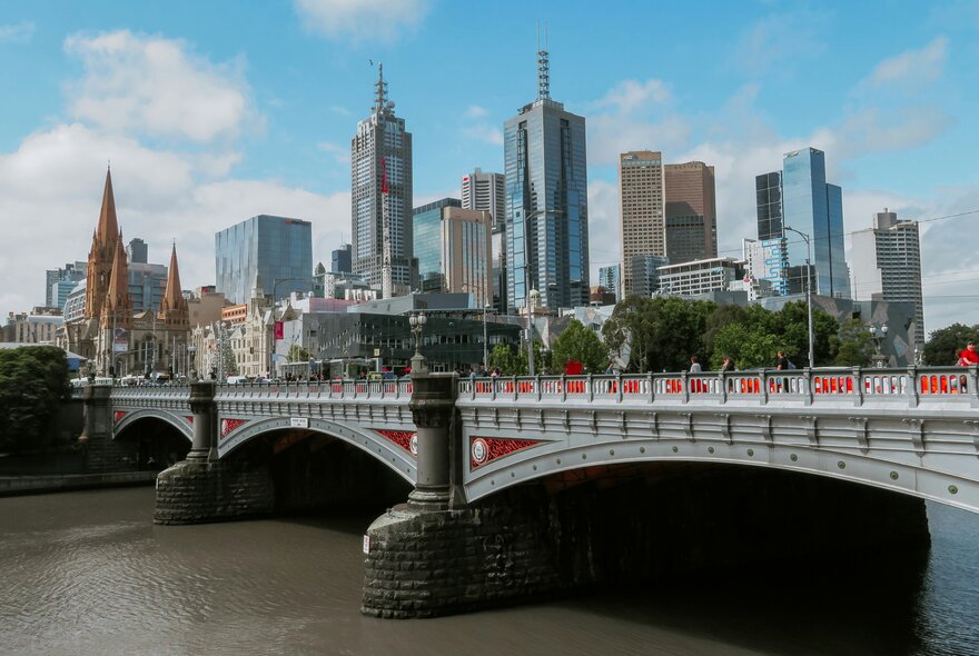 Melbourne city building skyline against a blue sky dotted with fluffy clouds, with Princes Bridge and the Yarra River in the foreground.