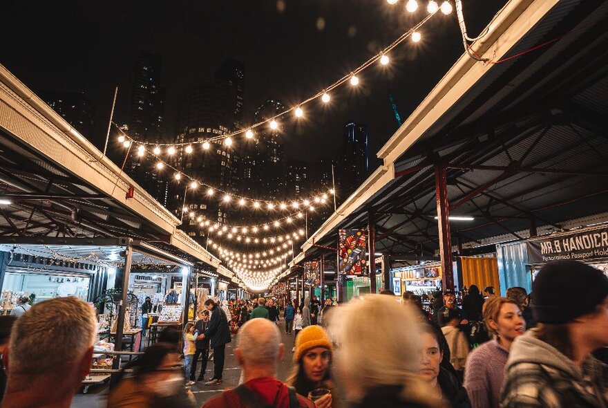 People walking around the Queen Victoria Market at twilight, the outdoor shed decorated with festive overhead lights.