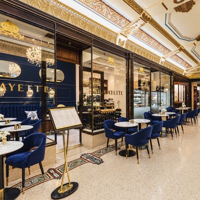 Outdoor tables and chairs outside a cafe with deep blue velvet seating and white marble tables set within an ornate, gold-trimmed historic arcade.