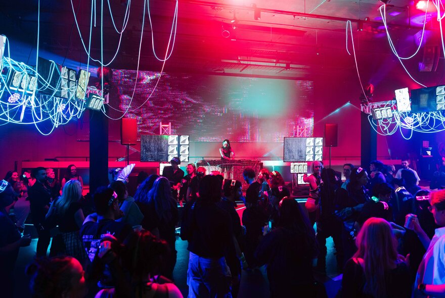 A crowded indoor venue filled with people dancing under vibrant red and blue stage lighting, with a DJ on a stage in the background.