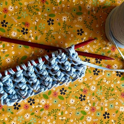 A small portion of knitting, in a blue yarn, with dark red knitting needles, displayed next to a ball of yarn and on a floral surface.