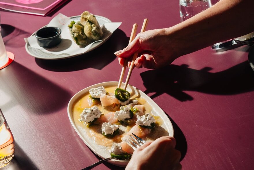 Hands using chopsticks to pick up food from a share plate of entrees on a purple table.