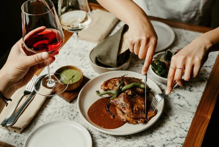 Someone cutting into a meat dish in a restaurant while another diner holds a glass of red wine.