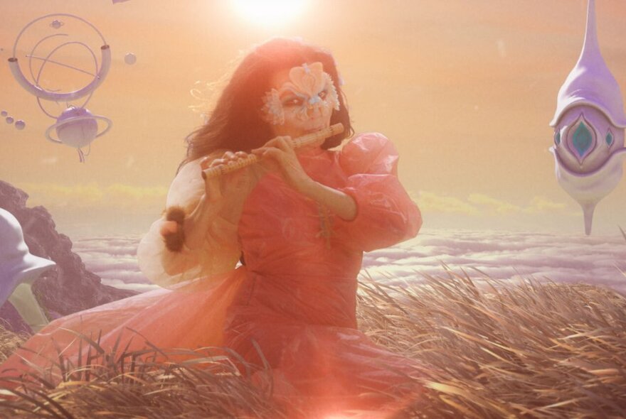 Singer and artist Bjork wearing a pink dress and playing a flute in a dreamy sci-fi landscape; a still taken from her music video for The Gate.