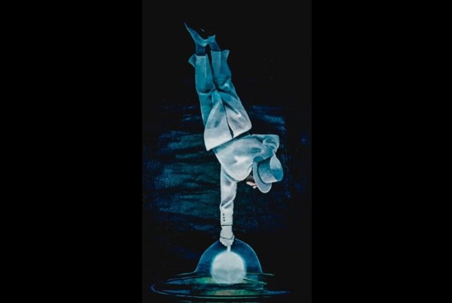 Magician balancing one finger on a globe, suspended updside down, wearing a white suit and hat.