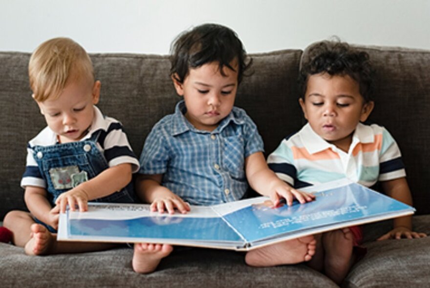 Three children seated on a couch reading a big picture book.